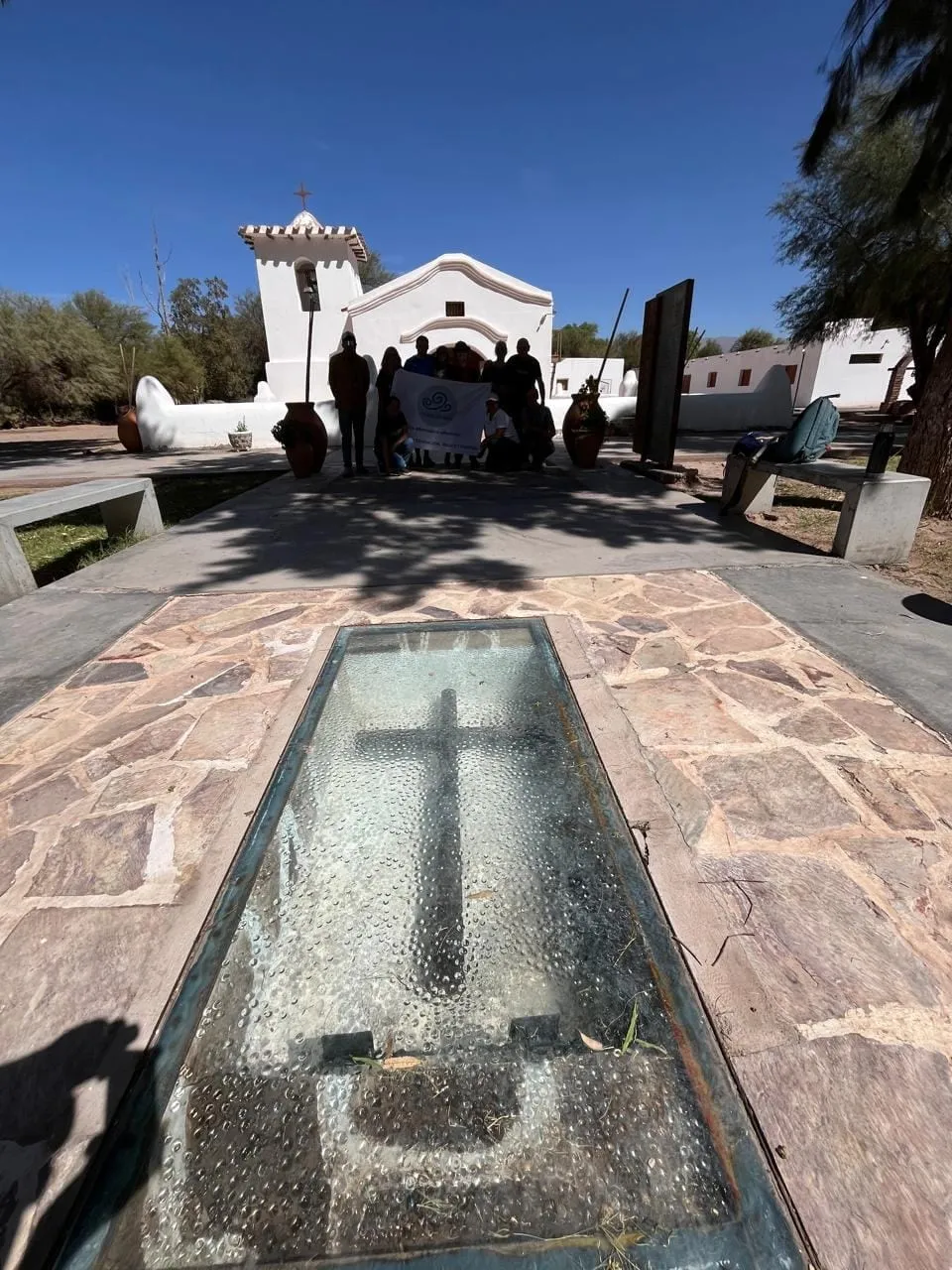 Una cruz reflejada en un espejo de agua de piedra, con una iglesia blanca al fondo y visitantes a lo lejos