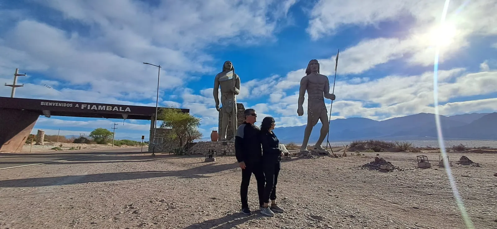 Dos grandes estatuas de figuras con lanza junto al cartel de bienvenida a Fiambalá, con una pareja delante