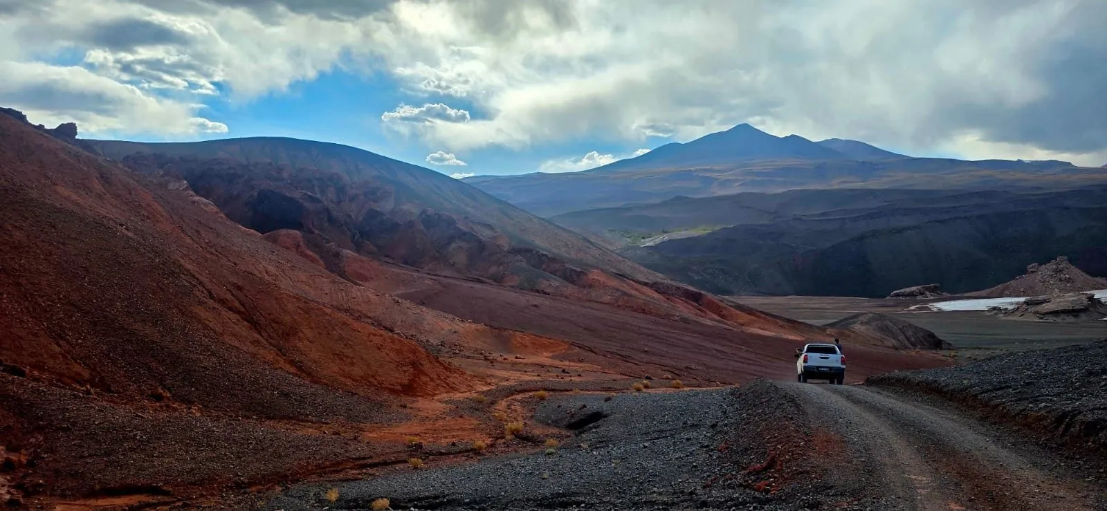Camino de tierra entre cerros rojizos y negros en la puna de Antofagasta de la Sierra, con una camioneta a lo lejos