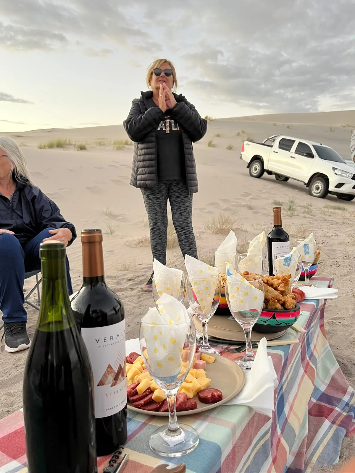 Mesa armada sobre la arena con botellas de vino, copas y una tabla de fiambres y quesos, con una camioneta blanca detrás