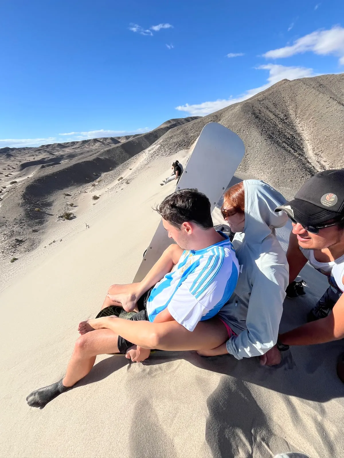 Viajeros sentados en la cresta de una duna con una tabla de sandboard