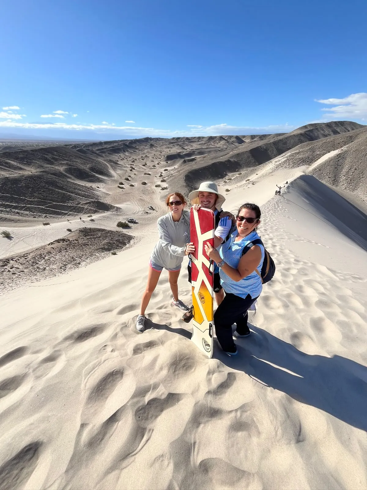 Tres viajeros posando sobre una duna con una tabla de sandboard