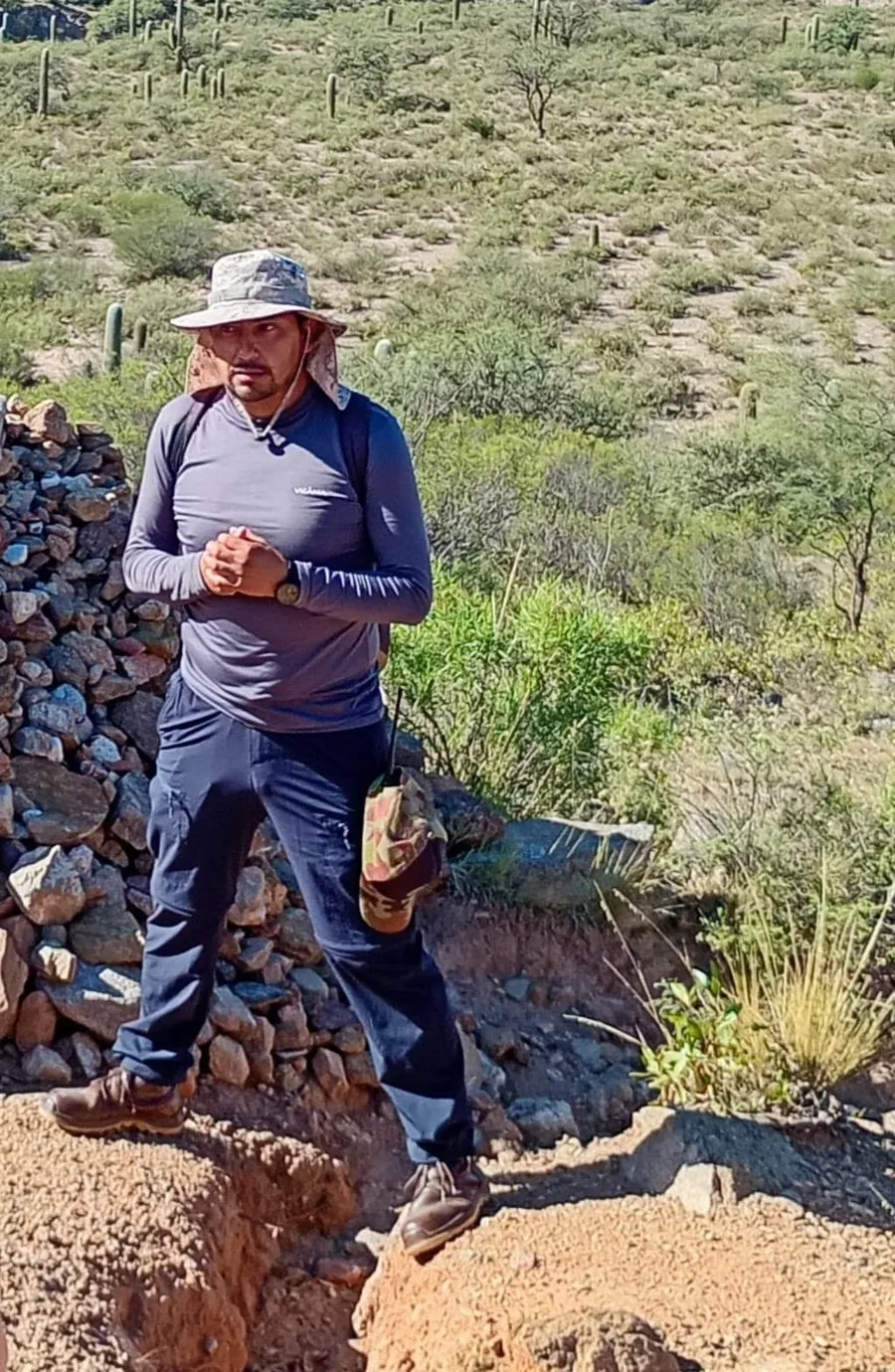 Dulio, guía local de los Castillos de Villa Vil, con sombrero y mochila entre cardones y un muro de piedra