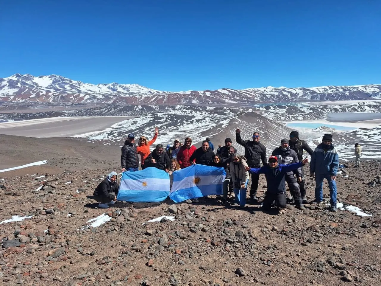 Un grupo de viajeros con banderas argentinas sobre un cerro con nieve y una laguna blanca detrás
