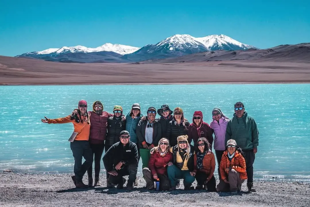 Un grupo de viajeros posando de rodillas y de pie junto a una laguna turquesa, con volcanes con nieve al fondo