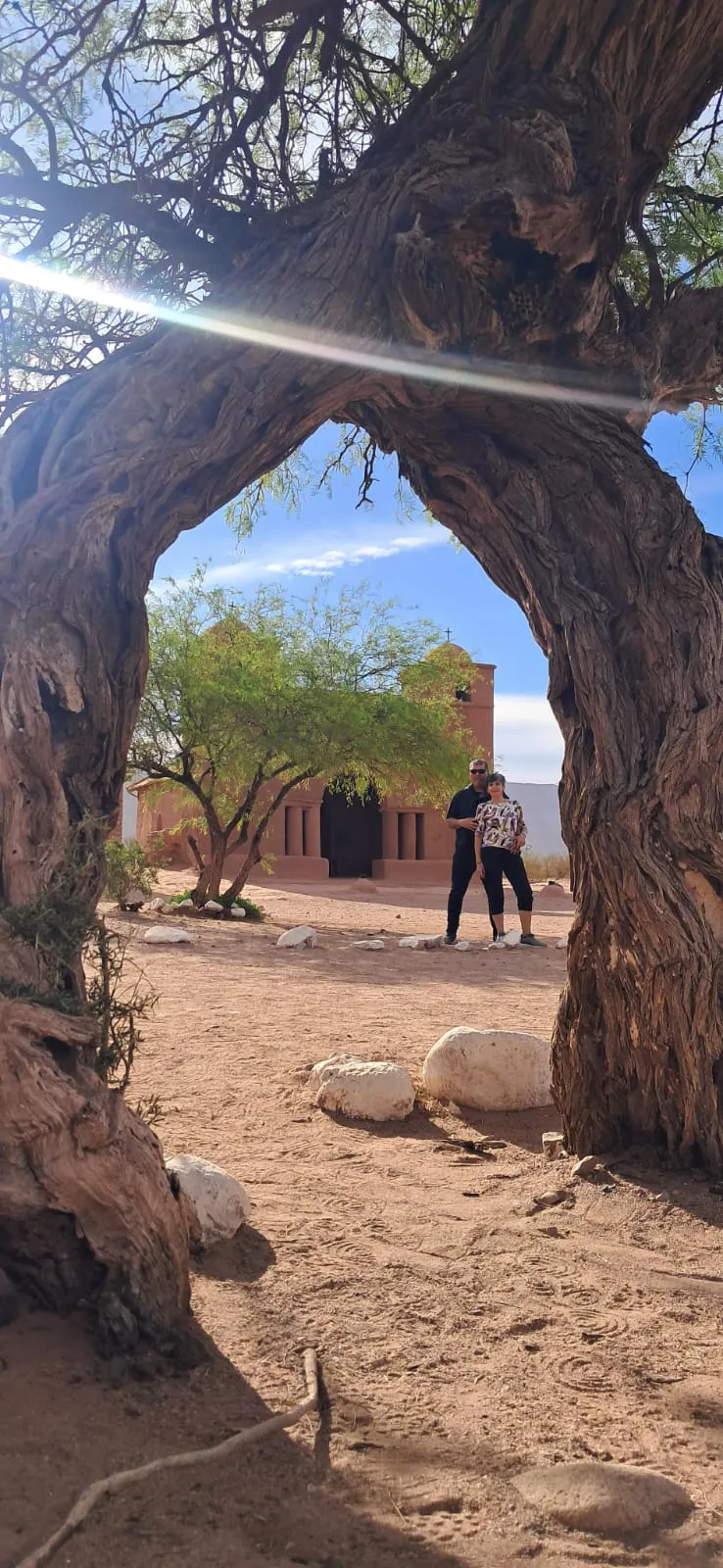 La capilla de adobe de Nuestra Señora del Rosario vista a través del arco de un algarrobo de tronco retorcido, con una pareja de visitantes