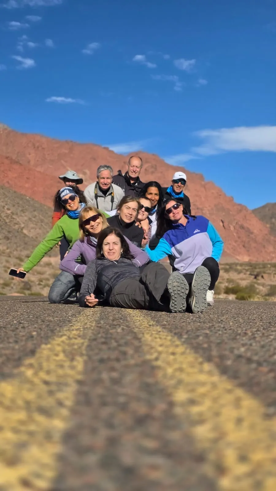 Un grupo de viajeros posando recostados sobre la ruta, con montañas rojas detrás