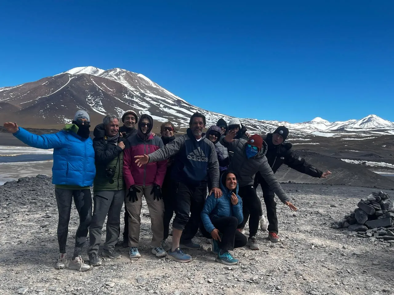 Un grupo de viajeros abrigados con los brazos abiertos frente a un volcán con nieve