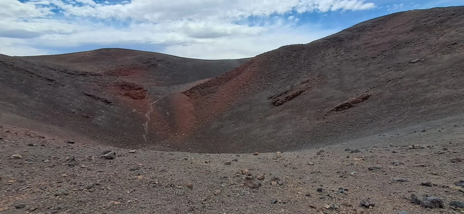 Borde de un cráter de tierra rojiza y marrón bajo un cielo con nubes