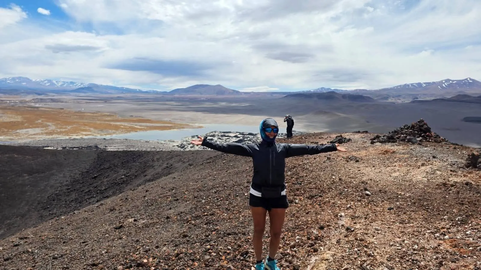 Una viajera con los brazos abiertos en una loma del volcán, con una laguna y cerros al fondo